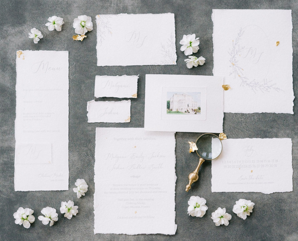 Flatlay fond gris d'un ensemble de papeterie de mariage avec loupe ancienne et fleurs blanches mis en scène par la wedding planner Amélie Pichon Weddings